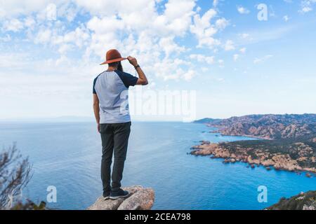 Rückansicht des bärtigen Mannes im Hut, der am Meer auf Stein steht und wegschaut. Stockfoto