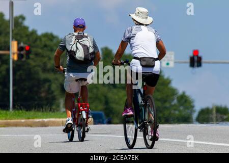 Ein Seniorenpaar trägt lässige Shorts und T-Shirts sowie Hüte fährt an einem sonnigen Sommertag Fahrräder in der Nähe einer Autobahn. Man hat ein faltbares Pedal Fahrrad Stockfoto