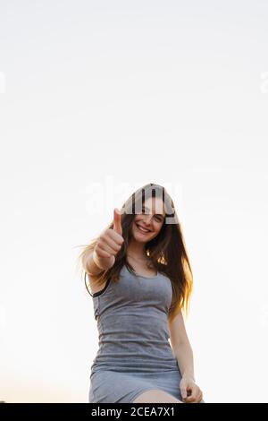 Wunderschöne junge Brünette weiblich stehen im Freien in lässigen Sommerkleid Und zeigt Daumen nach oben Geste Blick auf die Kamera und lächelt Auf hintergrundbeleuchtetem Hintergrund des klaren Himmels von unten Stockfoto