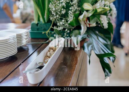 Schmale weiße Platte mit Snack und Löffel auf Holz serviert Tisch mit Tellern und blühenden Blumen Stockfoto