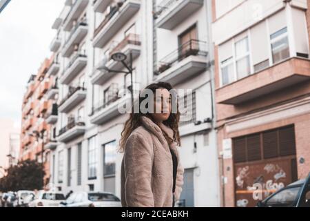 Schöne junge Frau mit lockigen Haaren, die in der Nähe eines modernen Gebäudes auf der Straße der Stadt auf die Kamera schaut Stockfoto