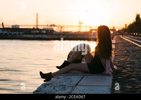 Seitenansicht Attraktive leidenschaftliche Dame auf Böschung in der Nähe von Wasser sitzen Oberfläche mit Schiff bei Sonnenuntergang Stockfoto