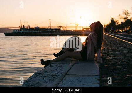 Seitenansicht Attraktive leidenschaftliche Dame auf Böschung in der Nähe von Wasser sitzen Oberfläche mit Schiff bei Sonnenuntergang Stockfoto