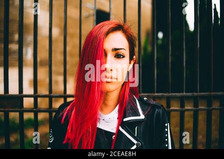 Schöne junge Frau in Lederjacke und mit gefärbten Haaren in roter Farbe Blick auf die Kamera Stockfoto