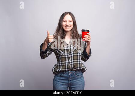 Kaffee mitnehmen. Glückliche junge Frau zeigt Daumen nach oben und hält Tasse Kaffee. Stockfoto