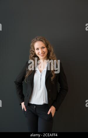 Schöne blonde junge Frau im dunklen eleganten Anzug, selbstbewusst mit gekreuzten Armen stehend und auf die Kamera lächelnd auf schwarzem Hintergrund schauend Stockfoto