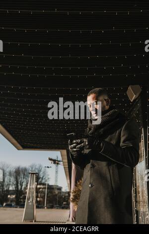 Von unten Aufnahme von schönen afroamerikanischen Mann mit modernen Smartphone während stehen in der Nähe von modernen Gebäude Dekoration für Weihnachten mit Feenleichte Girlanden Stockfoto