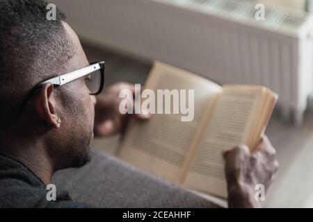 Erwachsene afroamerikanische Mann in Brille Buch lesen, während sitzen Auf dem Sofa zu Hause Stockfoto