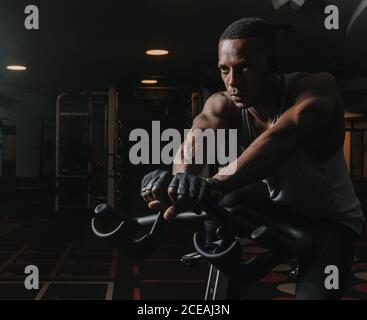 Schöner afroamerikanischer Mann sitzt auf Heimtrainer während des Trainings Im dunklen Fitnessraum Stockfoto