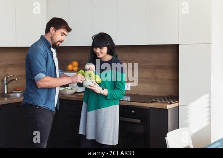 Junger Mann und Frau in lässigen Outfits lächeln und halten Schüssel mit frischem Salat beim Kochen in der modernen Küche zusammen Stockfoto