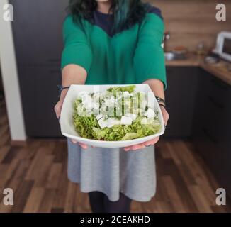 Crop weiblich in lässigen Kleid Demonstration Schüssel mit frischem Salat Zur Kamera, während Sie in der stilvollen Küche zu Hause stehen Stockfoto