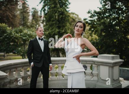 Charmante junge Frau in schönen Hochzeitskleid posiert für Kamera, während in der Nähe Bräutigam auf der Terrasse des Luxus-Gebäude stehen Stockfoto