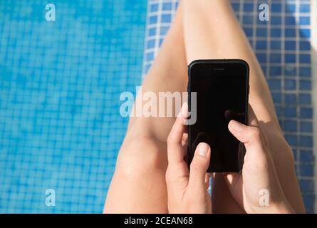 Von oben Schuss von anonymen weiblichen Browsing Smartphone mit leer Bildschirm beim Sitzen in der Nähe des Swimmingpools mit sauberem Wasser Stockfoto