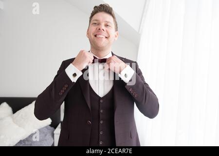 Junger gutaussehender lächelnder Mann in schwarzem Kostüm im Schlafzimmer stehend Und das Anziehen der Fliege Stockfoto