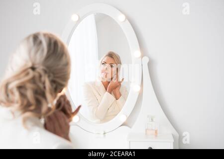 Rückansicht einer jungen, schönen Frau in weißem Kostüm mit Frisur, die im Zimmer vor dem Frisiertisch steht und im Spiegel schaut Stockfoto