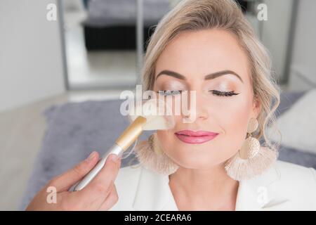 Crop-Ansicht der Person, die Make-up für junge schöne Frau im Schlafzimmer mit geschlossenen Augen und lächelnd sitzen Stockfoto