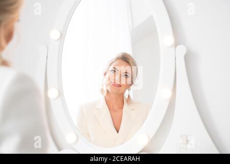 Rückansicht einer jungen, schönen Frau in weißem Kostüm mit Frisur, die im Zimmer vor dem Frisiertisch steht und im Spiegel schaut Stockfoto