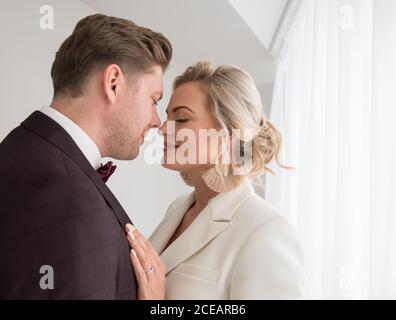Junger, hübscher Mann in schwarzem Kostüm und eine schöne Frau in weißer Jacke, die im Zimmer neben dem Fenster steht und umarmt Stockfoto