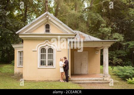 Seitenansicht der eleganten Braut und Bräutigam umarmen in der Nähe klein Altes Haus im grünen Garten Stockfoto