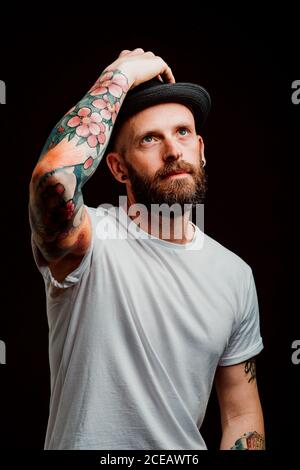 Fröhliche bärtigen Hipster in Hut und t-shirt mit Tattoos auf Arme auf schwarzem Hintergrund Stockfoto