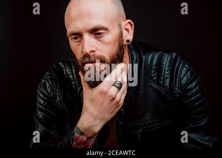 Nachdenklich kahlen Hipster in Leder Jacke mit Tattoos auf Hand kratzen Bart auf schwarzem Hintergrund Stockfoto