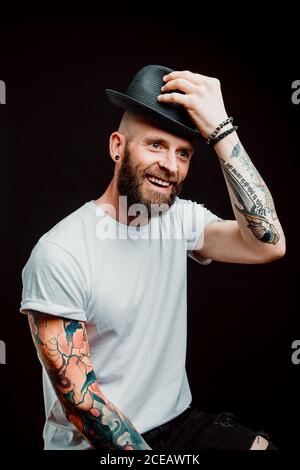 Fröhliche bärtigen Hipster in Hut und t-shirt mit Tattoos auf Arme auf schwarzem Hintergrund Stockfoto
