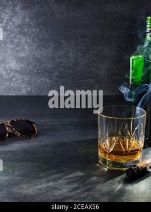 Komposition aus Kristallglas mit goldenem Whiskey auf grauem Hintergrund Mit Sonnenbrille und Zigarre rauchen Stockfoto