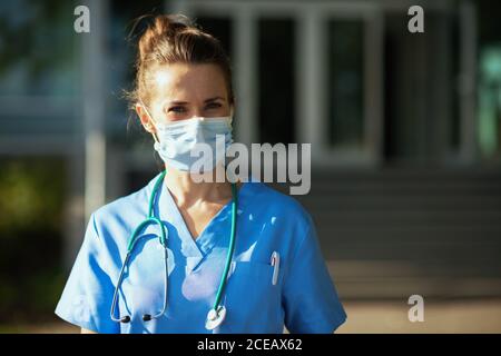 Covid-19 Pandemie. Portrait der lächelnden modernen Arzt Frau in Uniform mit Stethoskop und medizinische Maske im Freien in der Nähe von Krankenhaus. Stockfoto