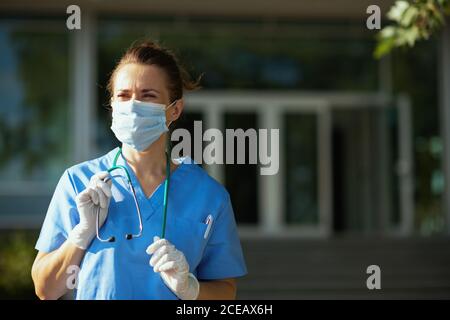 Coronavirus Pandemie. Zuversichtlich moderne Ärztin in Uniform mit Stethoskop und medizinische Maske Blick in die Ferne in der Nähe von Krankenhaus. Stockfoto