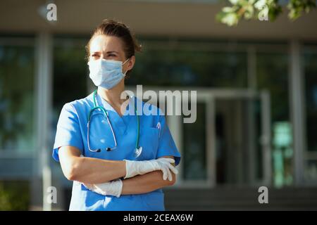 Covid-19 Pandemie. Zuversichtlich moderne Ärztin in Uniform mit Stethoskop und medizinische Maske Blick in die Entfernung außerhalb des Krankenhauses. Stockfoto