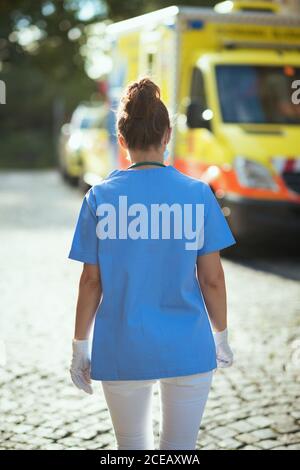 Coronavirus-Pandemie. Von hinten gesehen Sanitäterin Frau in Scrubs mit medizinischer Maske zu Fuß außerhalb in der Nähe von Ambulanz. Stockfoto