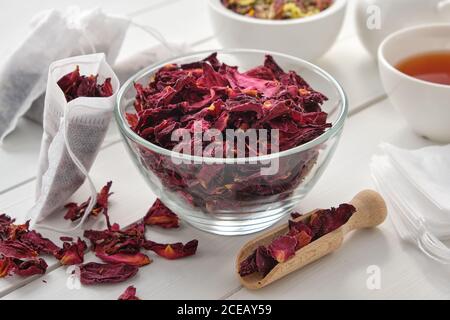 Schüssel mit trockenen Rosenblättern, Teebeutel mit roten Blütenblättern der Heidrose gefüllt. Kräuterteetasse, Teekanne und Mörser auf Holztisch. Stockfoto