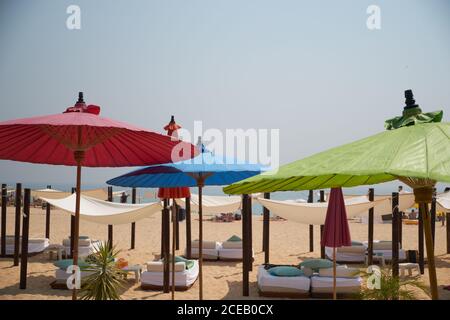 Bunte Holzmöbel und Sonnenschirme am Sandstrand mit Gründbeet Laub auf dem Hintergrund mit klarem Himmel und endlosem Ozean Stockfoto