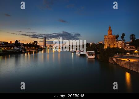 Ruhiger, glatter Fluss mit Booten, die in der Nähe der Küste schwimmen Ufer der Nacht Stadt von Laternen auf dem Hintergrund beleuchtet Blauer wolkig Himmel Stockfoto