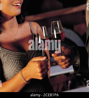Ansicht einer jungen Frau in Kleid und eines Mannes in Kostüm, der im Zimmer steht und Rotwein aus Gläsern trinkt Stockfoto