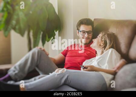 Fröhlicher Mann und Frau liegen und sitzen auf dem Sofa, halten die Fernbedienung und Fernsehen Stockfoto