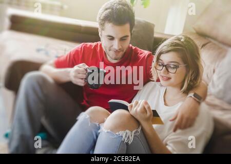 Mann, der Kaffee trinkt, während er mit seiner Freundin auf dem Boden sitzt und ein Buch liest Stockfoto