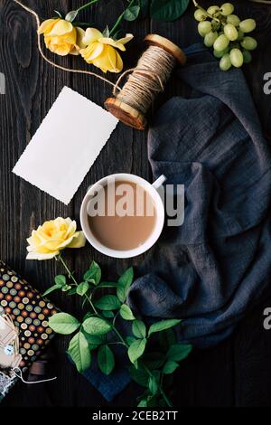 Von oben Schöne gelbe Rosen und leerer Hinweis in der Nähe von Tasse frischen Kaffee auf Holz Tischplatte inmitten Stück blauen Tuch mit Leinenfaden und Ri liegen Stockfoto