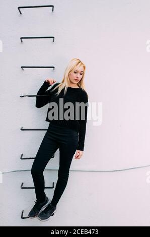 Junge hübsche blonde Frau, die an der weißen Wand auf einer Leiter hält. Stockfoto