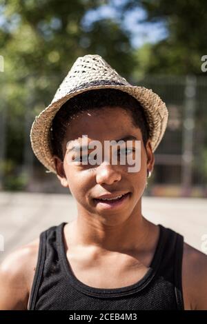 Afro Junge posiert mit Strohhut auf dem Basketball gericht seiner Nachbarschaft und lächelt Stockfoto