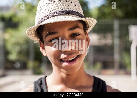 Afro Junge posiert mit Strohhut auf dem Basketball gericht seiner Nachbarschaft und lächelt Stockfoto