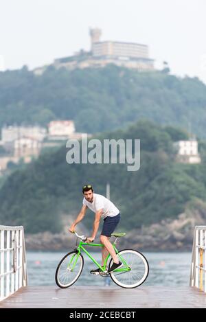 Junger sportlicher Mann, der Fahrrad auf dem Pier am Meer reitet Die Stadt Stockfoto