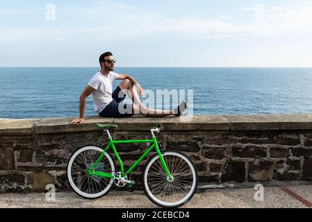 Junger Mann in Sonnenbrille sitzt an der Uferwand und entspannt Auf dem Fahrrad Stockfoto