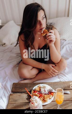 Junge Frau, die auf dem Bett appetitliche Waffeln und leckere Getränke isst Stockfoto