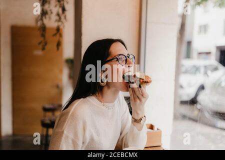 Seitenansicht der hübschen jungen Frau im stilvollen Outfit beißen Leckeres Sandwich, während man in einem gemütlichen Café neben dem Fenster sitzt Stockfoto