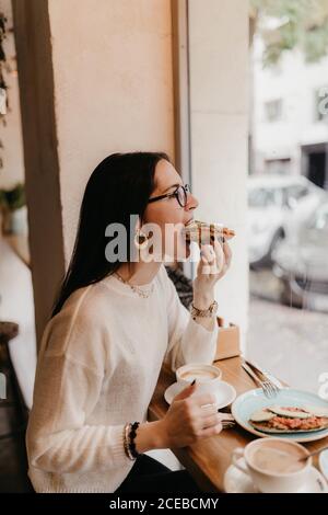 Seitenansicht der hübschen jungen Frau im stilvollen Outfit beißen Leckeres Sandwich, während man in einem gemütlichen Café neben dem Fenster sitzt Stockfoto