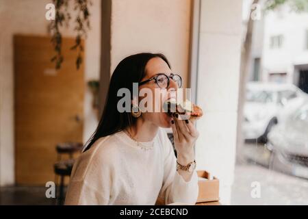 Seitenansicht der hübschen jungen Frau im stilvollen Outfit beißen Leckeres Sandwich, während man in einem gemütlichen Café neben dem Fenster sitzt Stockfoto