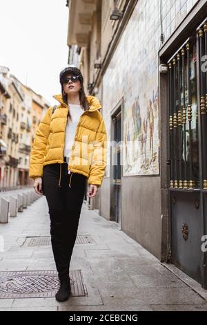 Schöne junge Frau in trendigen Outfit und Sonnenbrille zu Fuß auf Bürgersteig in der Nähe eines wunderschönen Gebäudes an der Stadtstraße Stockfoto