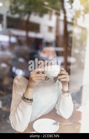 Schöne junge Frau in trendigen Outfit hält Tasse heiß Trinken und wegschauen, während Sie hinter dem Glas sitzen Gemütliches Café Stockfoto