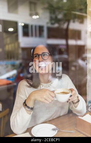 Schöne junge Frau in trendigen Outfit hält Tasse heiß Trinken und wegschauen, während Sie hinter dem Glas sitzen Gemütliches Café Stockfoto